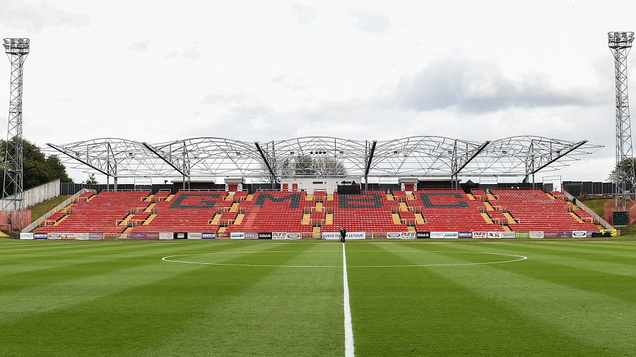 A picture showing the grass pitch at Gateshead International Stadium. The picture is taken from the half-way line looking across the pitch. On the other side is a stand full of red seats. A number of black seats spells out GMBC.