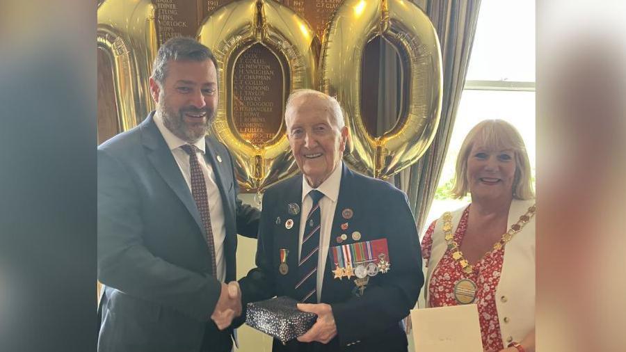 Image depicting councillor Kevin Guy in a navy suit and red tie, shaking hands with D-Day Veteran Stan Ford who is wearing his military regalia and holding a gift received from the council. Also pictured is councillor Karen Walker who is wearing a white waistcoat, red and white top and white trousers.
