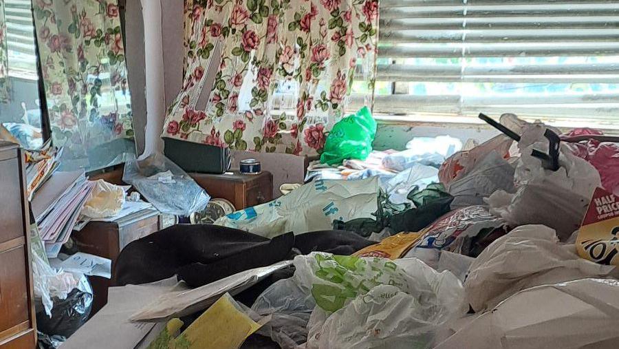 A bedroom full of plastic bags and paperwork. Sun is shining through the window, with a floral curtain hanging over blinds. 