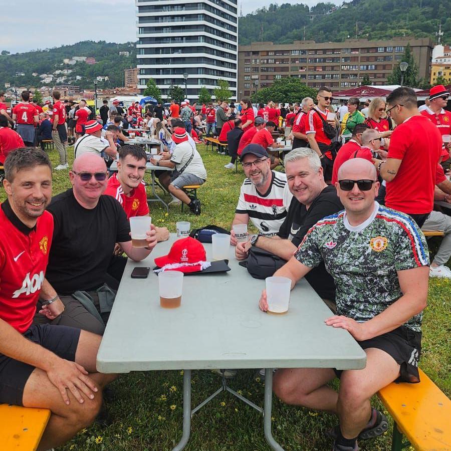 Five men, including Jamie and Allan sitting on a bench in a park in Bilbao. The group have cups of beer. A large high rise building is behind them.