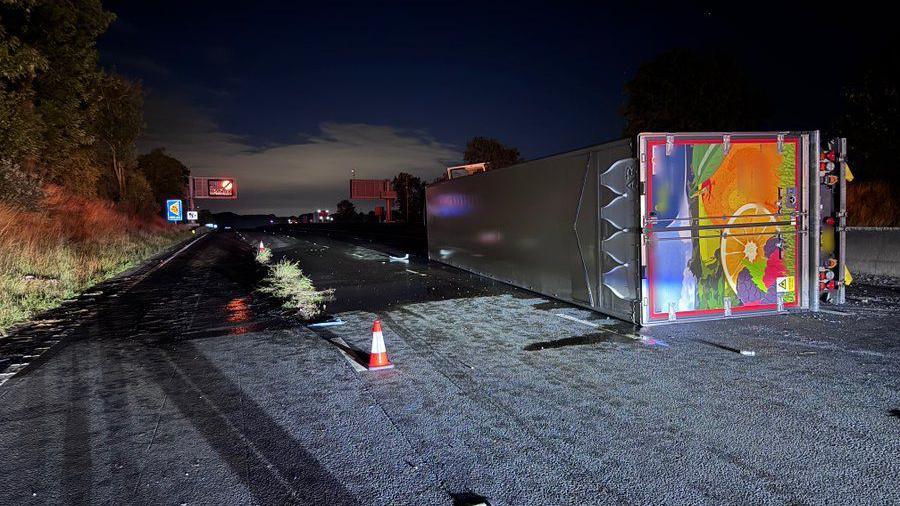 A lorry overturned on the first lane of a motorway at night. The lorry lies on its left side, illuminated by an unknown source of light. It has a silver roof and a picture of fruit on its back. An illuminated gantry in the distance is showing a closure sign. A grassy bank can be seen to the left.