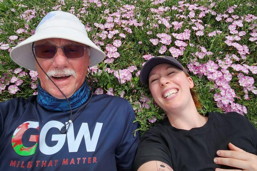 John and Kyla smile for a selfie as they lay in a flower bed for a rest during their walking challenge. The purple flowers surround their heads, they both wear sunhats. 