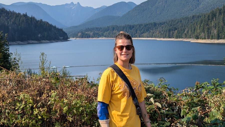 Amy wearing a yellow t-shirt and stood by a lake with mountains in the background