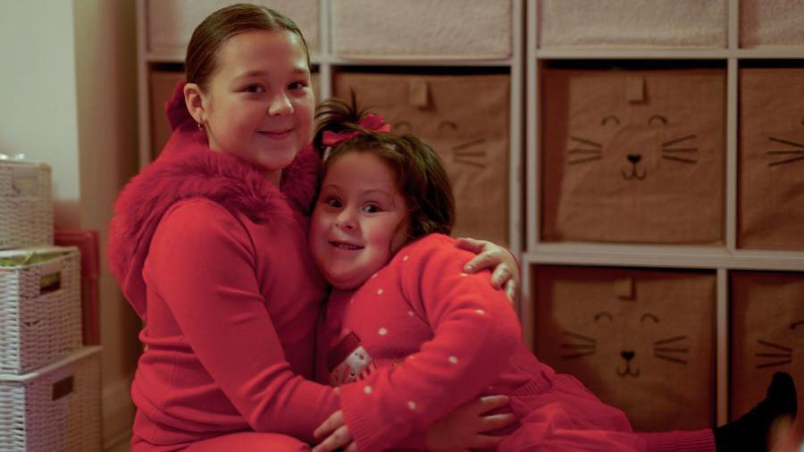 Amber and Laila hugging each other while looking into the camera. Both are wearing red clothes for Christmas, with a snowman on Amber's top.