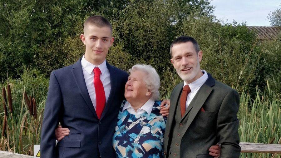 Will Radford is on the left with short dark hair wearing a navy blue suit with a white shirt and red tie. In the middle, his grandmother laughs as she looks up at him. She has white curly hair and wears a patterned jumper over a white shirt. On the right Will's father has greying hair and wears a grey suit and waistcoat with a white shirt and brown tie.