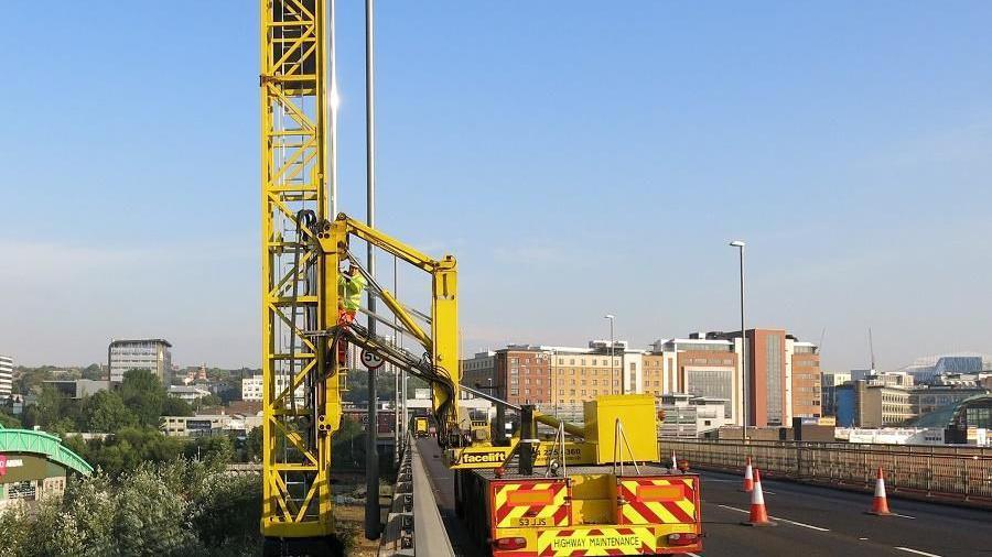 A highway maintenance vehicle parked on the Redheugh Bridge between Newcastle and Gateshead. The bright yellow vehicle holds a large piece of equipment which is stretched over the side of the bridge. The equipment holds a large ladder which is standing vertical off the side of the bridge. A man in a hi-vis jacket is climbing on to the ladder.