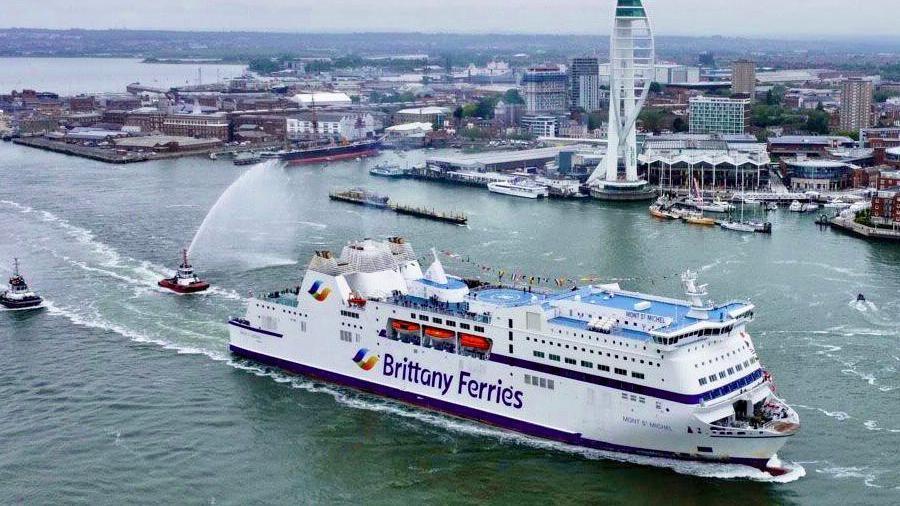 The Barfleur leaving Portsmouth Harbour with tugs following behind and the shoreline of Portsmouth in the background on an overcast day