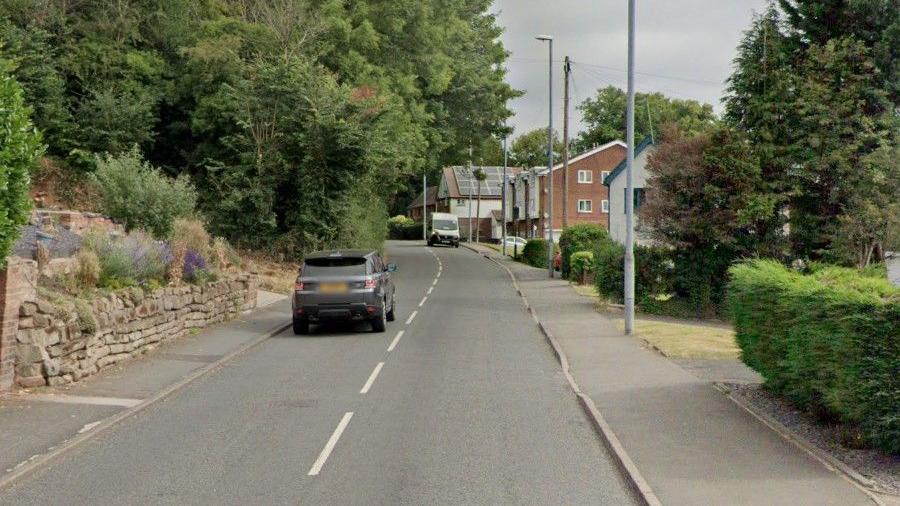 A stretch of B-road through a village, with a stone wall in front of a house on one side, trimmed hedges on the other, and cars on the road.  