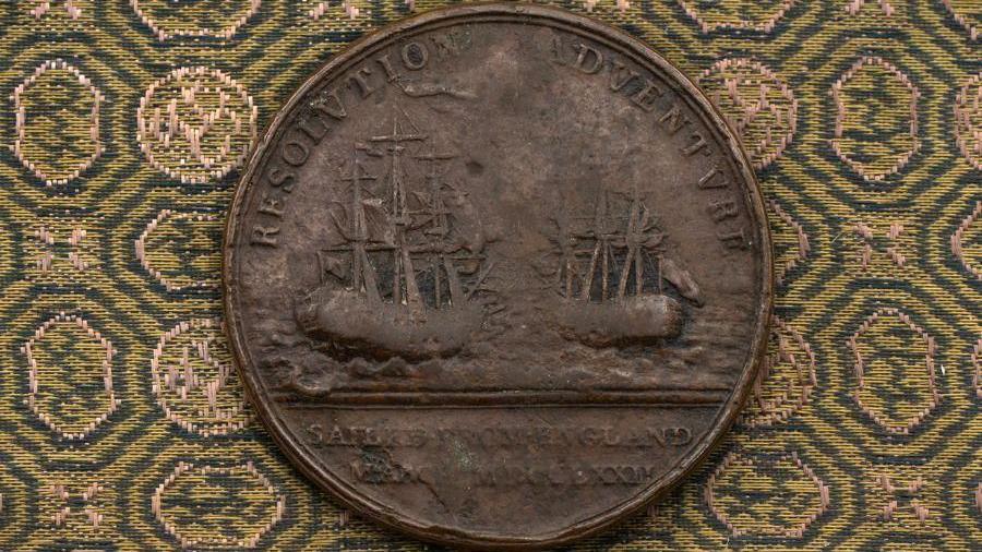 A medal with images of the ships used by James Cook on his second voyage. The medal is pleased on a green and yellow patterned cloth.