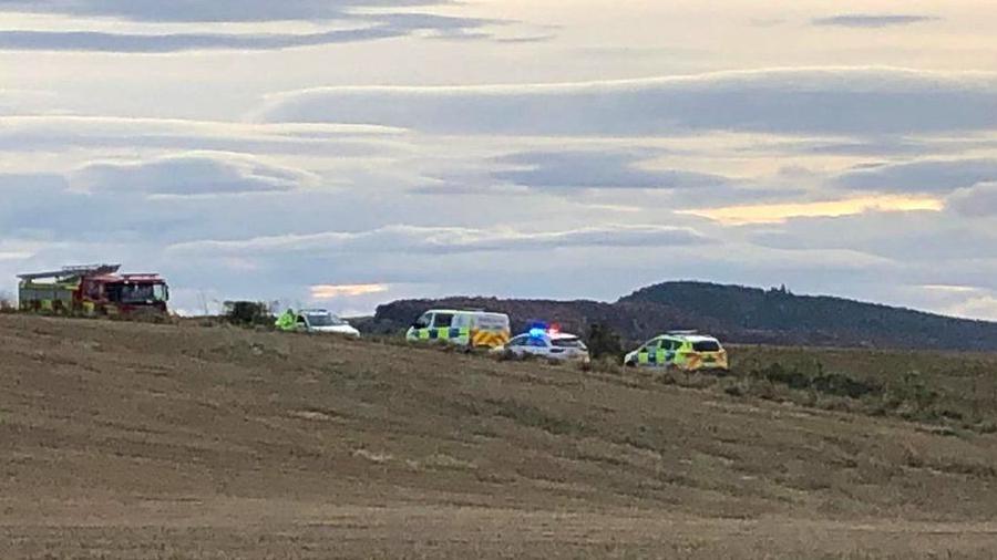 A fire engine and other emergency vehicles, including police cars are parked near fields. One of the vehicles' emergency lights are flashing.