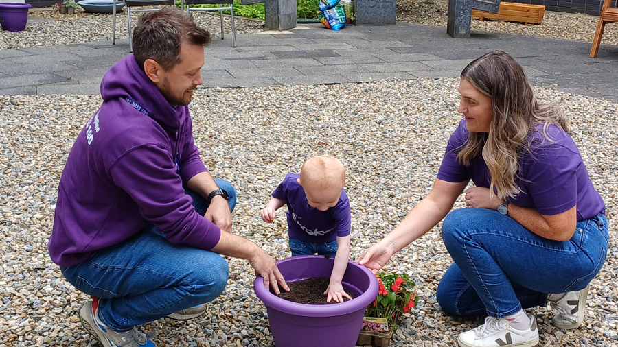 Shows a man on the left, children in the middle and woman on the right, all wearing purple tops and playing with a flower pot.