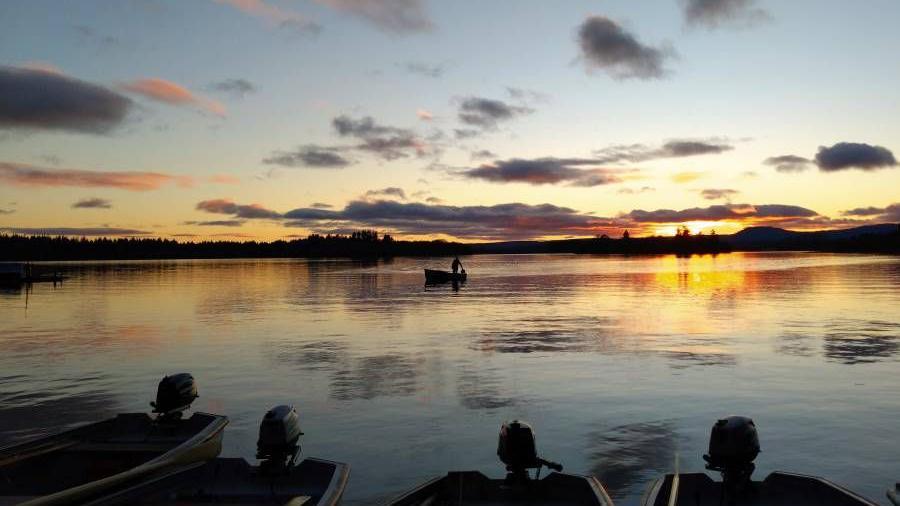 Rowboats rest on a still lake as the sun sets behind distant hills.