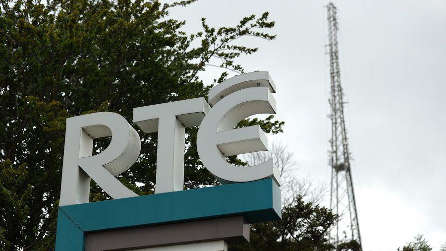 An RTE sign outside the broadcaster's building in Dublin. The sign has a green base and white letters and is raised off the ground. A pylon is in the background.