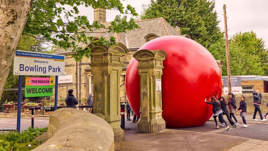 A large red inflatable ball is pictured being pushed through a stone pillar entrance at Bowling Park in Bradford by a group of four young girls.