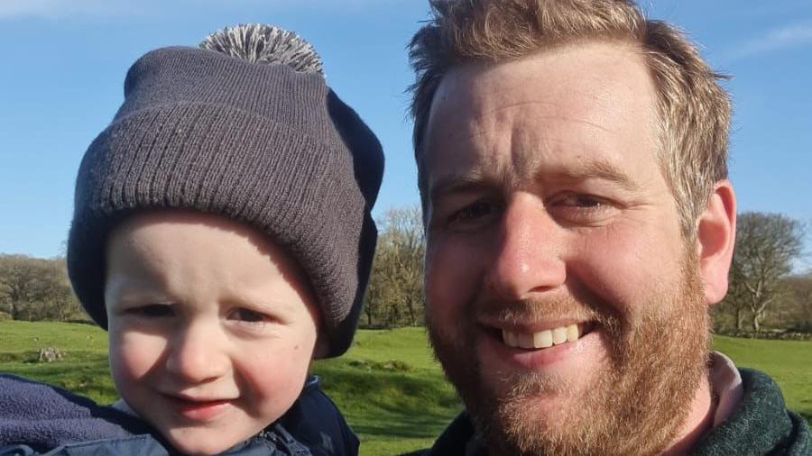 Bearded farmer Neil Austin smiles while holding his son on their farm in Galloway.