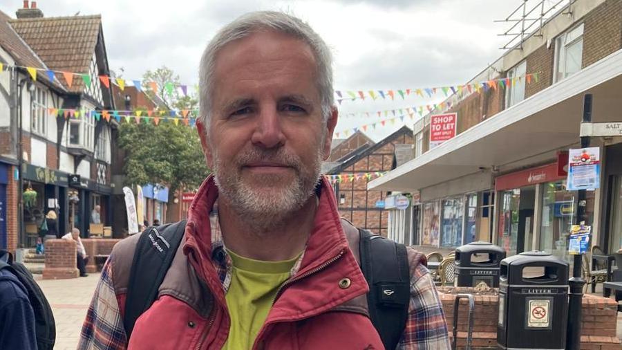 Paul Esling, a resident of Northwich, in the town centre. He has short grey hair, a trimmed beard, and wears a lime green t-shirt with a plaid shirt and red jacket.