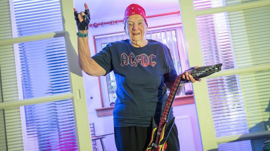 A woman dressed for a rock concert by AC/DC - she has a band T-shirt on and a bandana, is throwing the devil's horns rock gesture and a inflatable guitar in her other hand
