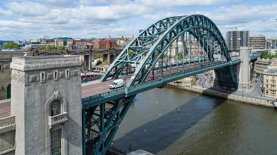 The Tyne Bridge which over the River Tyne. It is a green, arched structure but the paint looks patchy and rusted. There are four white stone towers, two on either side of the river. The High Level Bridge can be seen behind it and various buildings in Newcastle.