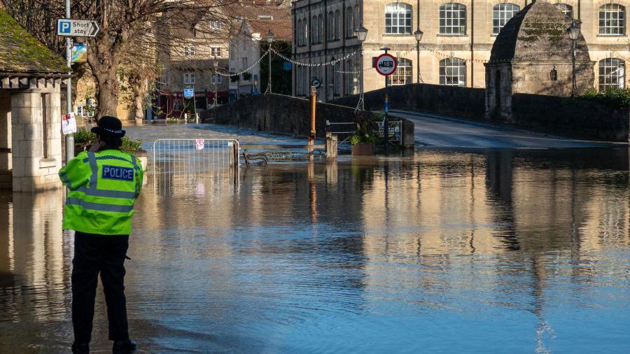 A police officer wearing a high-vis jacket and hat stands by a flooded road next to a bridge after the River Avon burst its banks
