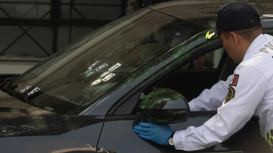 A police officer wearing blue plastic gloves and a white uniform shirt examines the steering wheel of the car of the mayor's private secretary. Bullet holes can be seen in the car's windscreen. Signs with numbers have been attached to the screen to mark the entry holes.