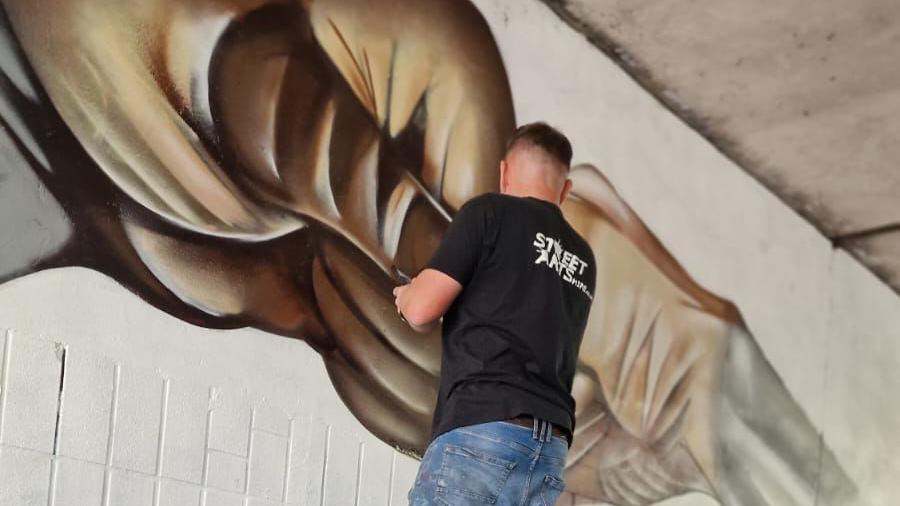 Nathan Murdoch stands with his back to the camera while painting a mural on a white wall. He is wearing a black T-shirt and blue jeans.