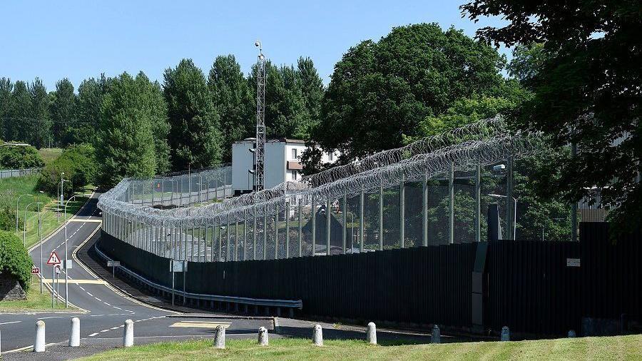 The perimeter fencing at Hydebank with a road running alongside it with trees throughout the site and a compound behind the wire 