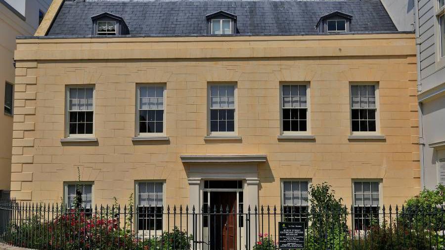 The photo shows the Georgian House. It is built from honey-coloured stone and has a door in the centre surrounded by windows. There is a black fence with spikes  in front of the house.