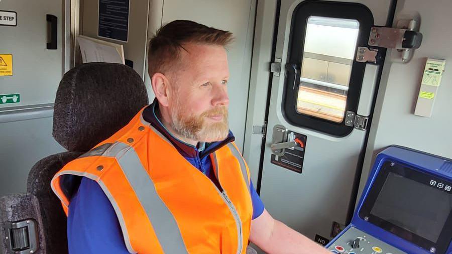 Dave Lay sitting in a train wearing an orange high vis jacket. He is concentrating on driving the train, looking out of the front window. He has short ginger hair and a full beard that is ginger flecked with some white.