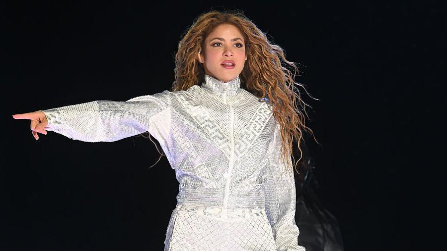 Shakira pointing in a silver-and-white top