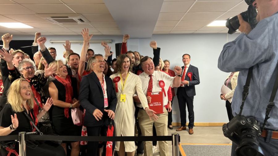 Jessica Toale wins Bournemouth West - she is wearing a white, fitted, long-sleeved dress and a red Labour rosette, and appears with other Labour Party members, all smiling broadly and at camera to the right of the frame