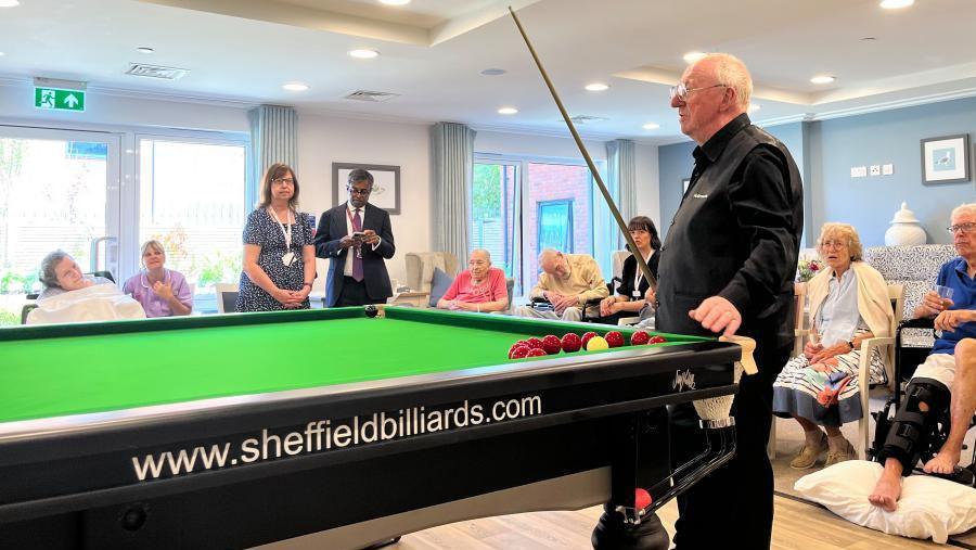 Dennis Taylor, standing at the end of a snooker table, with a cue pointing upwards. Behind him, there are care home residents.
