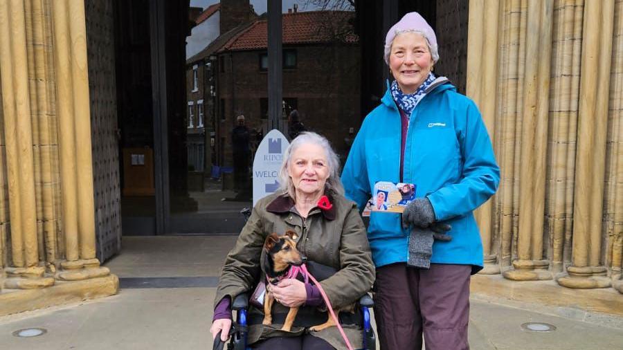 Two women and two dogs are gathered outside an entrance to a cathedral with tall, ornate columns and an arched doorway. One woman sits in a wheelchair, warmly dressed and holding a small dog in their lap. Beside them stands another woman, also bundled up, holding a leaflet.