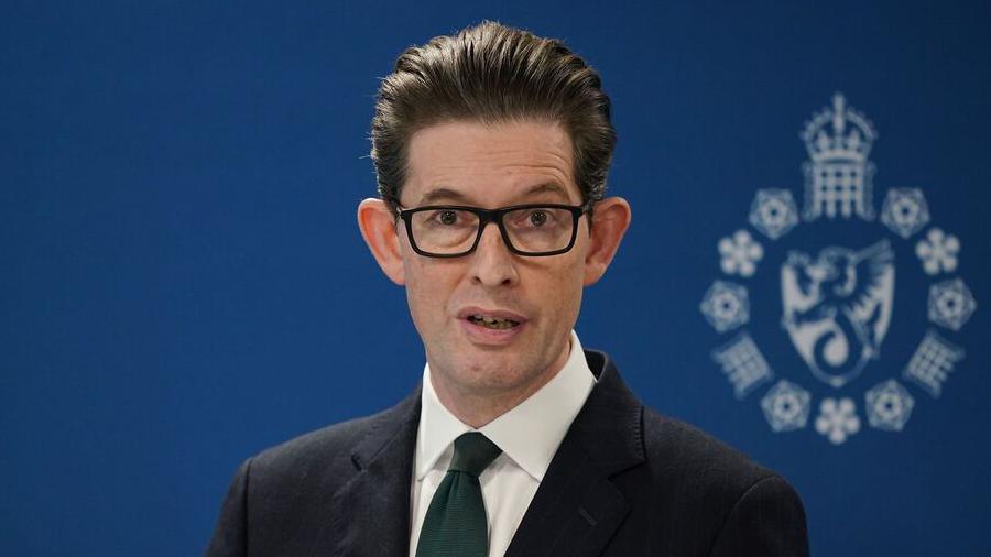 Ken McCallum wears a dark suit and glasses. He stands in front of a dark blue screen. 