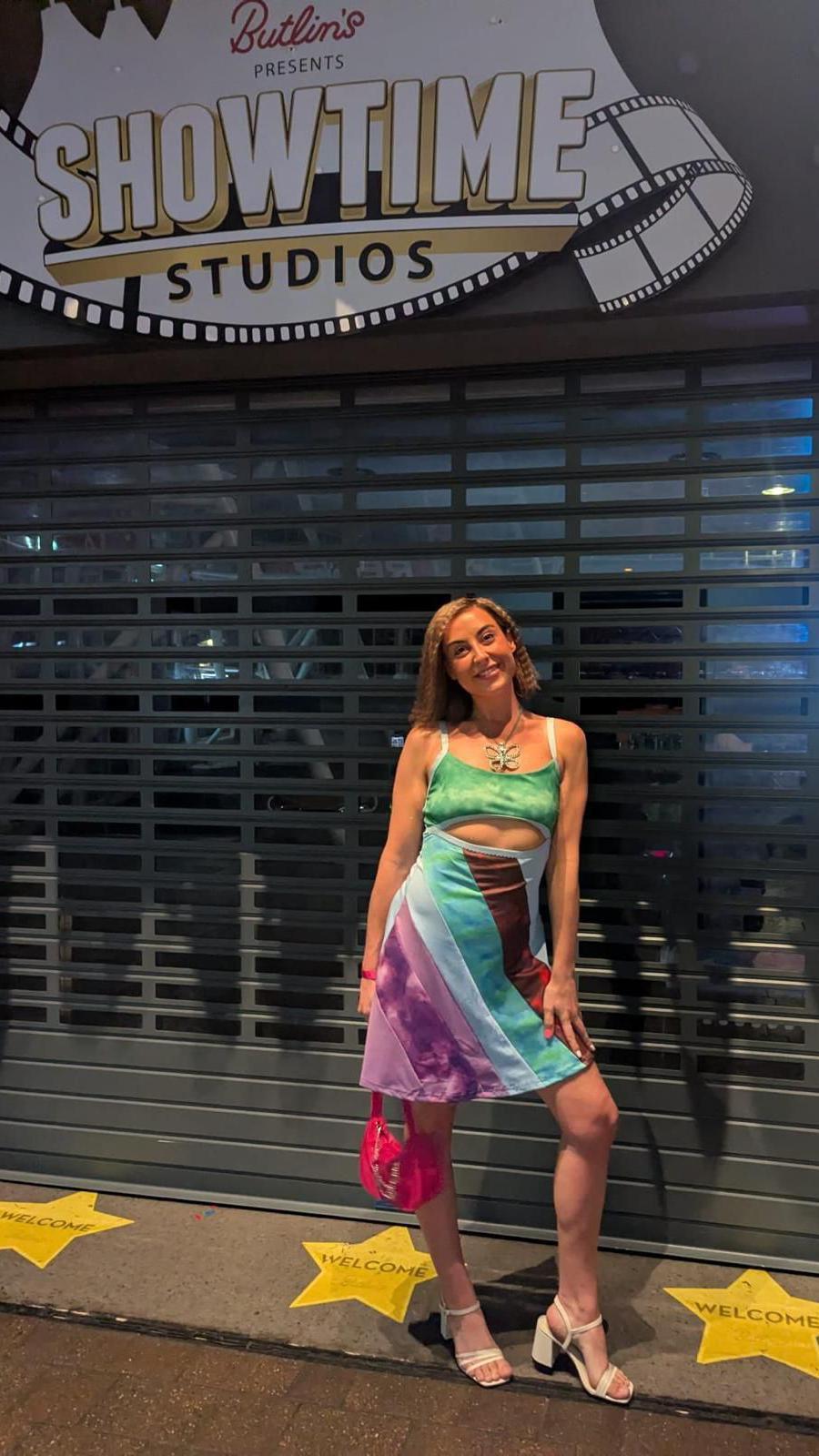 A woman wearing a multicoloured, striped skirt and green crop top, with white heels and a pink handbag. She's stood smiling, in front of a shutter, with yellow stars on the ground and a sign above her which reads: Butlin's presents showtime studios".