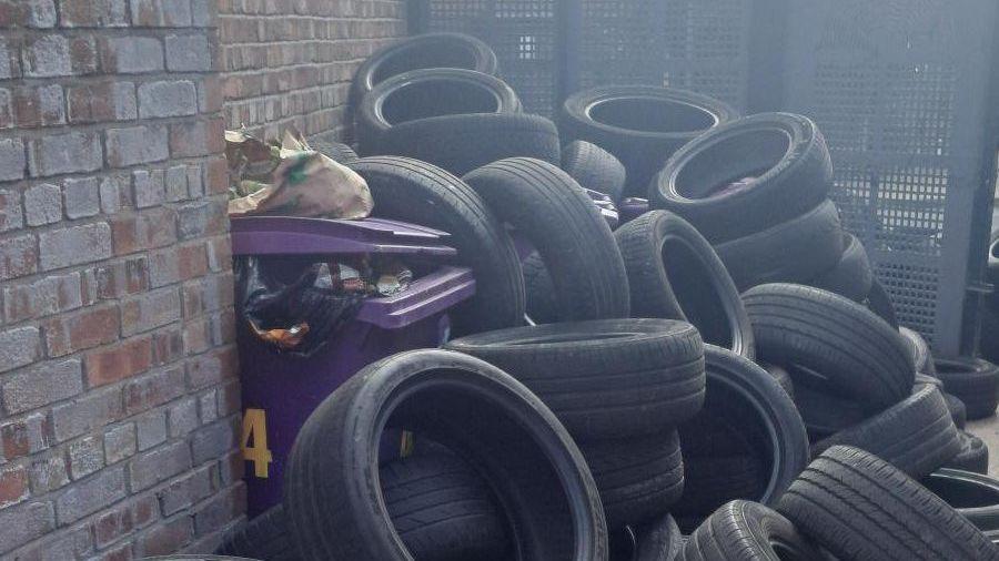 Dozens of black vehicle tyres piled beside metal alley gates between brick terraced houses.