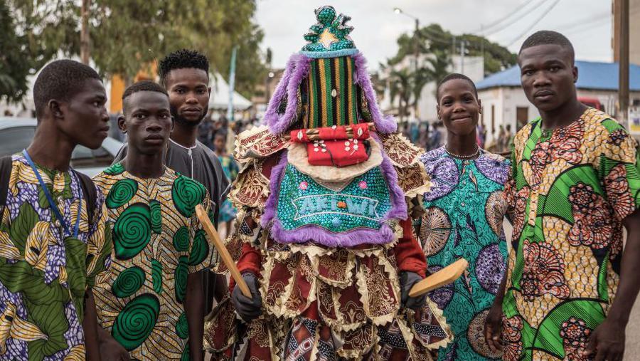 Visitors pose with an egungun, a character wearing a Yoruba costume and mask, during a festival in Porto-Novo, Benin - Sunday 4 August  a character wearing a Yoruba costume and mask during a festival in Porto-Novo, Benin - Sunday 4 August 2024