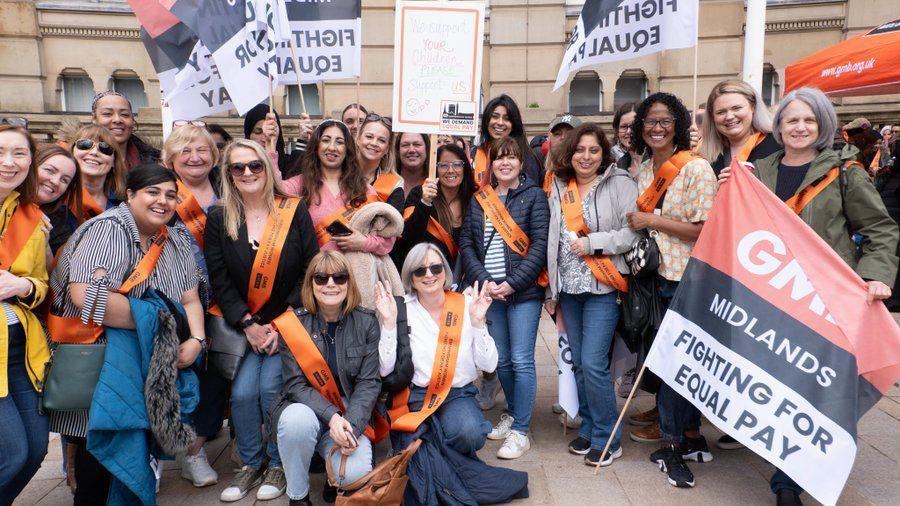 A large group of women outside a sandstone building. All those pictured are wearing orange sashes and a woman at the front holds a large flag which says "GMB Midlands Fighting For Equal Pay". Several other women at the back hold similar flags. Two of the women are crouching down at the front and all are smiling.