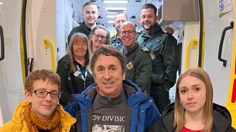 A family of three, from left to right, Mum, Dad and Daughter, standing in front of an ambulance crew of six people. They are all standing at the back of an ambulance which has its doors open so you can see into the vehicle. All nine people are smiling and looking into the camera.  