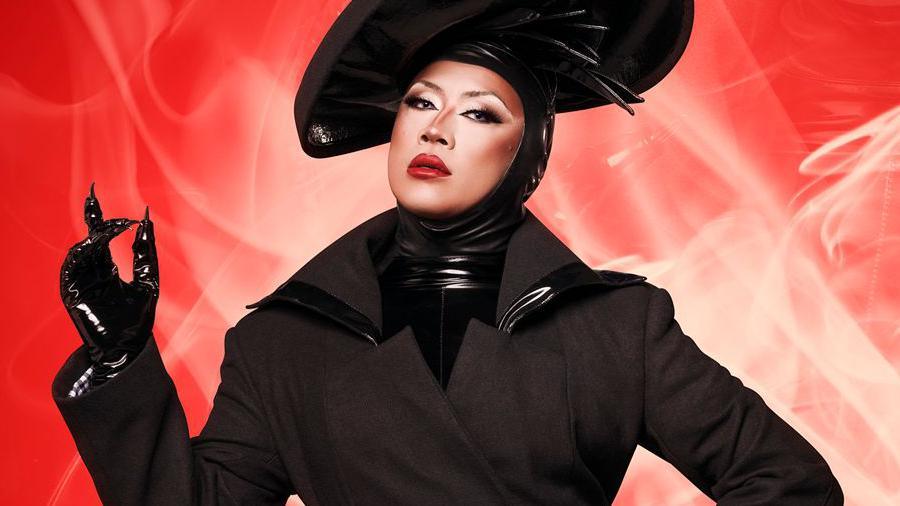 Wisps of white smoke surround Drag Race UK contestant Saki Yew who is wearing a dramatic, shiny black headdress that surrounds her face. She is covered in a thick, black coat with a winged collar and is wearing shiny black gloves with sharp fingertips. Her lipstick is red, as is the background she is in front of.