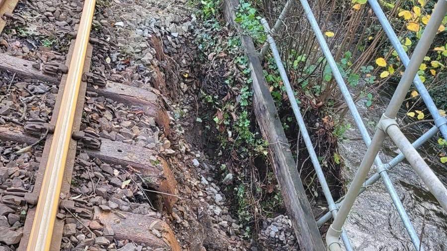 The side of a railway track which shows stones on the right crumbling down a bank. On the right is the bronze railway track. To the left of the fallen stones, there are bushes.