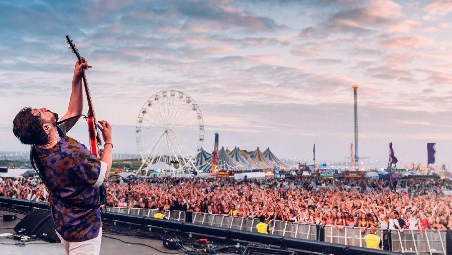 A musician holding a guitar and playing, there is a big ferris wheel and circus tops as well as a large audience watching the person on stage