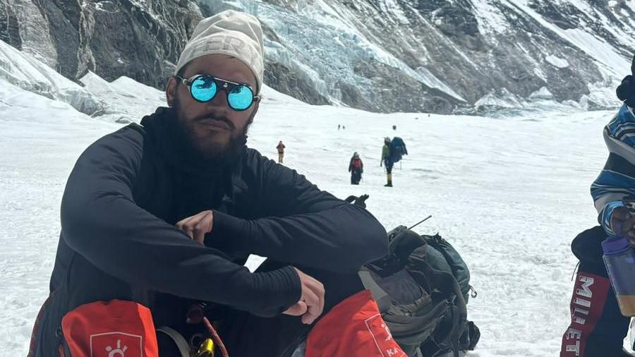 A man in mountain gear and sunglasses sitting on the ground with a mountain in the background