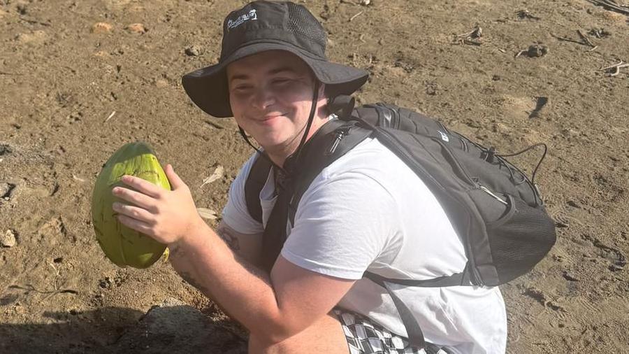 Ashton Taylor, with a black sun hat on, wearing a black rucksack is crouched down holding a large piece of fruit