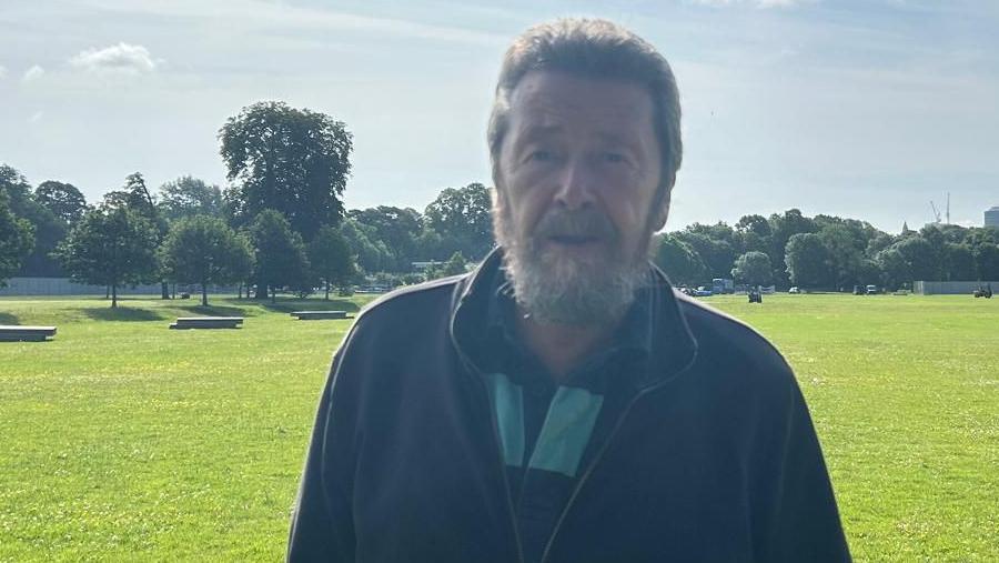 Llun o Paul Barret yn gwynebu'r camera gyda gwyrddni'r parc tu ol iddo