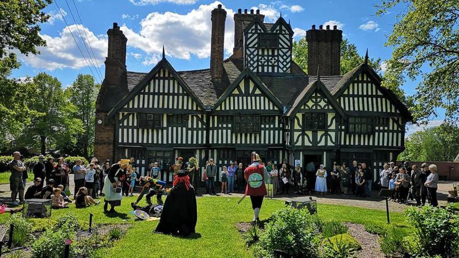 A large house from the 1600's with black horizontal and vertical markings. People dressed in medieval costumes are lined in front of the building. There is a green space to the front of the house with various bushes