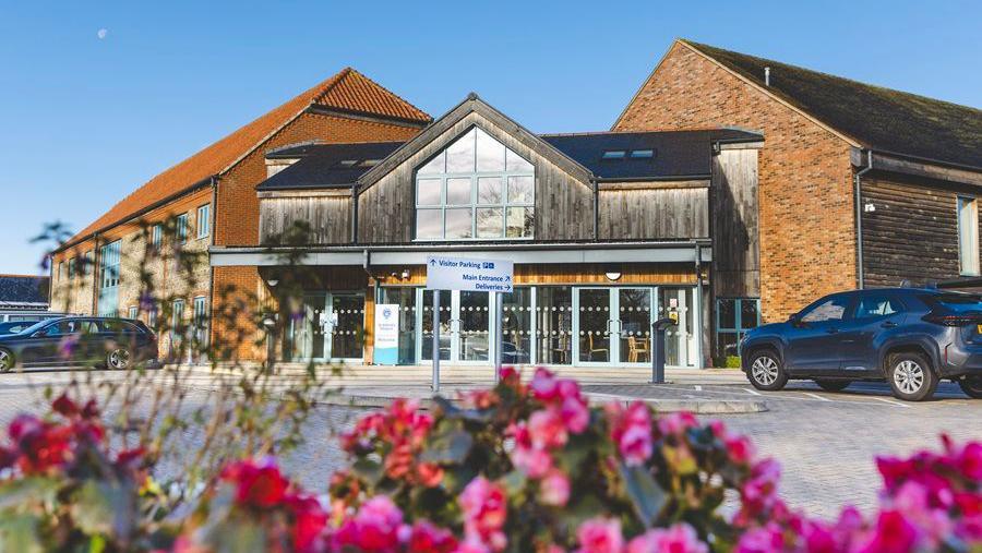 St Wilfrid's Hospice in Bosham, Chichester. There is a blue car parked outside the building, as well as some pink flowers in front. 