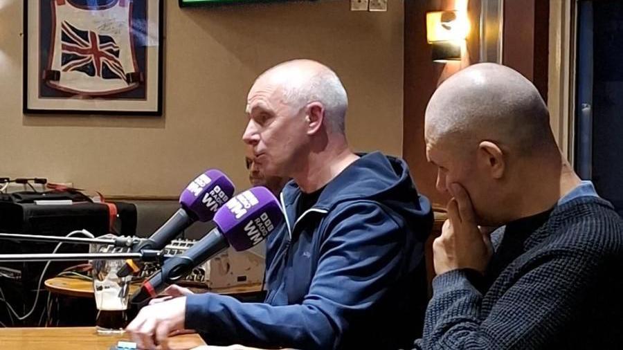 Two men sat a table with microphones inside a pub. Both men are bald and are wearing navy blue jumpers. 