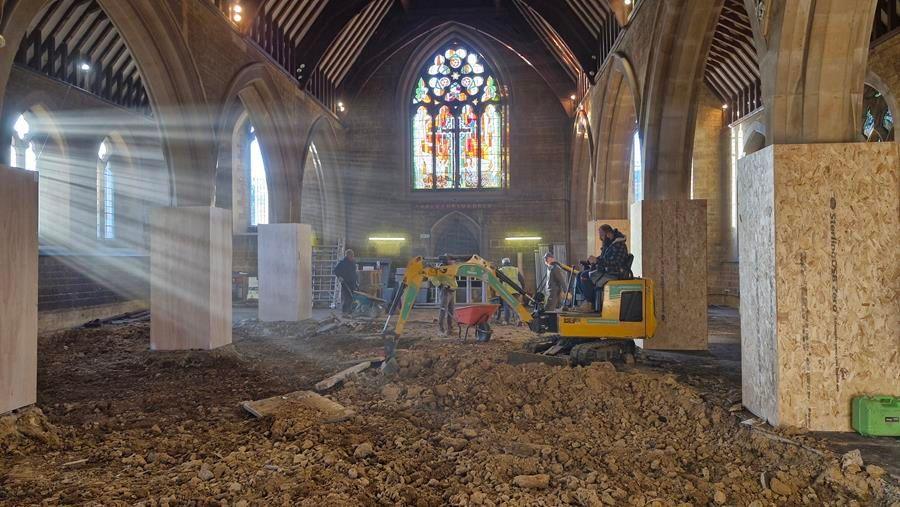 Church interior, showing stained glass window at the East End. The floor has been removed in the foreground, revealing soil beneath. A man is sitting on a yellow digger. Sun is streaming through the windows to the left