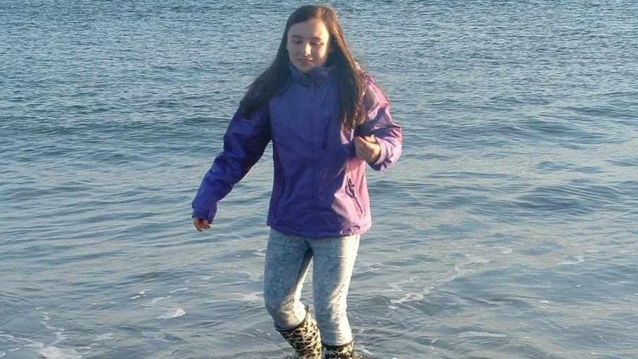 Rachel is walking on shallow sea water in black and white wellie boots. She is wearing denim jeans and a purple short coat. Her hair is long and brown. 