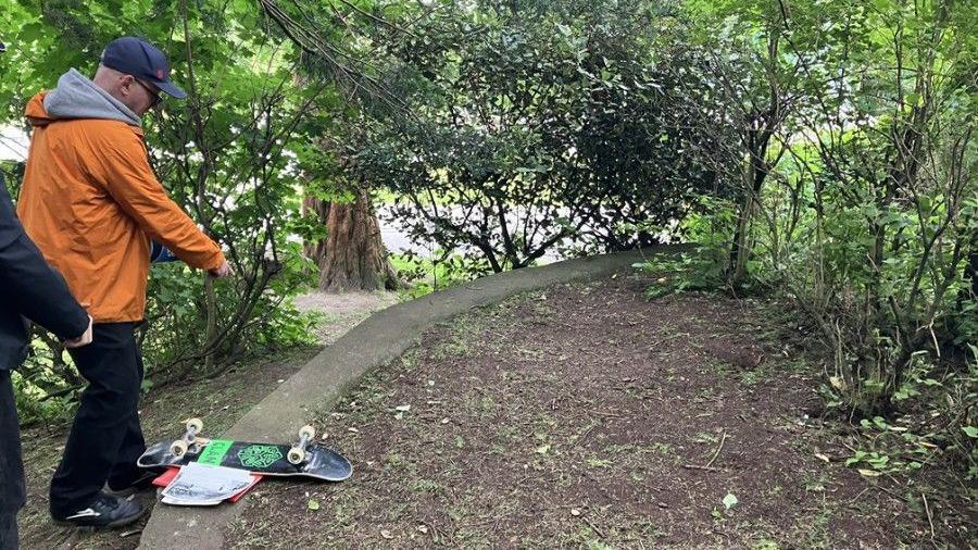 A man in an orange jacket and baseball cap pointing at an area of grass and dirt, where a skatepark used to be